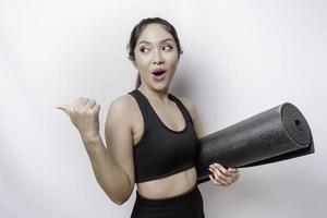 mulher asiática desportiva chocada vestindo roupas esportivas está segurando um tapete de ioga em fundo branco isolado. foto