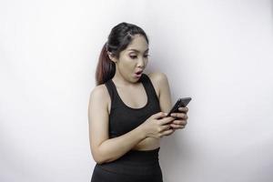 mulher asiática desportiva chocada vestindo roupas esportivas e segurando seu telefone, isolado por fundo branco foto
