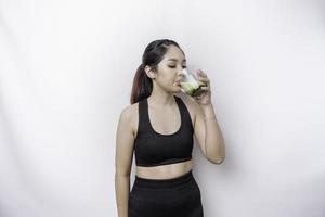 alegre mulher asiática esportiva vestindo roupas esportivas está bebendo um copo de saboroso smoothie verde, isolado no fundo branco. foto