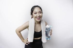 mulher asiática esportiva posando com uma toalha no ombro e segurando uma garrafa de água, sorrindo e relaxando após o treino foto