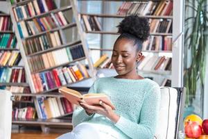 garota afro-americana lendo um livro sorrindo feliz e relaxada em um dia de lazer em casa. biblioteca em casa com estantes na parte de trás. foto