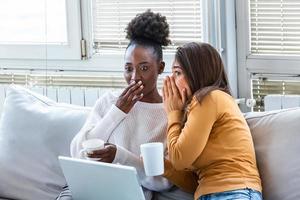 diversas amigas compartilhando segredos. duas mulheres fofocando em casa. garota emocional animada sussurrando no ouvido de sua amiga enquanto está sentada na sala de estar foto