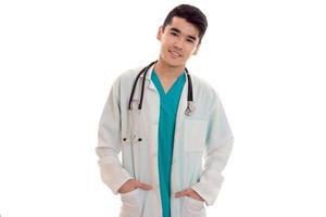 jovem médico bonito morena de uniforme com estetoscópio no pescoço posando na câmera isolada no fundo branco foto