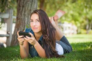 atraente, feliz, jovem, mestiça, mandando mensagens de texto em seu telefone celular do lado de fora, deitada na grama foto