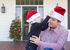 jovem pai e filha usando chapéus de Papai Noel na varanda da casa com decorações de natal foto