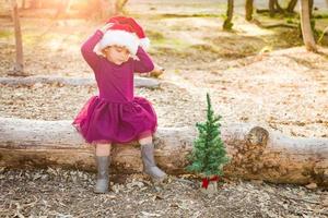 menina bonita de raça mista se divertindo com chapéu de papai noel e árvore de natal ao ar livre no log foto