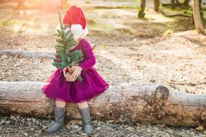 menina bonita de raça mista se divertindo com chapéu de papai noel e árvore de natal ao ar livre no log foto