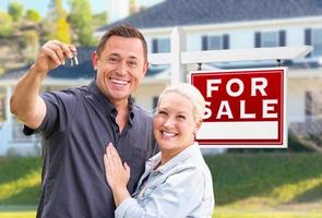 jovem casal adulto com as chaves da casa na frente de casa e à venda sinal imobiliário foto