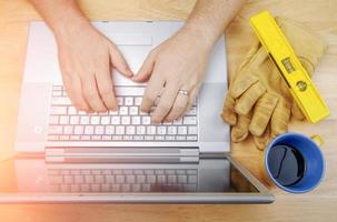 luvas de trabalho, nível e xícara de café ao lado do empreiteiro revisando o projeto no laptop foto