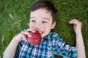 menino chinês e caucasiano de raça mista com maçã relaxando de costas na grama. foto
