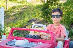 jovem menino chinês e caucasiano de raça mista usando óculos escuros brincando no carro de brinquedo foto