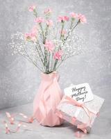 feliz dia das mães nota na caixa de presente com fita, cravo rosa, gypsophila branco em vaso em cinza. foto