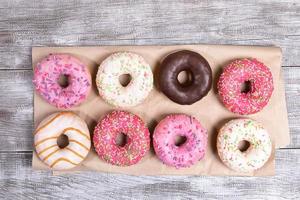 oito donuts tradicionais com esmalte multicolorido cuidadosamente dispostos em papel de embalagem na mesa de madeira pintada de branco. foto