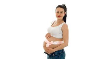 retrato de estúdio de uma jovem morena grávida com fita rosa na barriga sorrindo para a câmera isolada no fundo branco foto