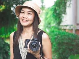 mulher asiática, usando chapéu e top preto sem mangas, de pé no jardim e segurando a câmera dslr, sorrindo alegremente. foto