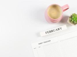 configuração plana do livro rastreador de hábitos, calendário de madeira de fevereiro, xícara rosa de café preto e vaso de plantas suculentas em fundo branco com espaço de cópia. foto