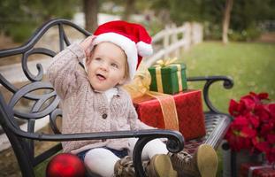 criança usando chapéu de Papai Noel sentado com presentes de Natal lá fora. foto