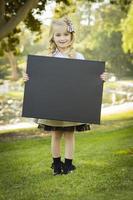 linda garotinha loira segurando uma lousa preta ao ar livre foto