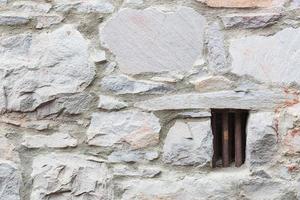 velho muro de pedra com pequena janela de cela de prisão com barras de ferro foto