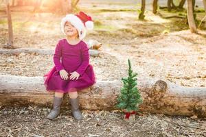 menina bonita de raça mista se divertindo com chapéu de papai noel e árvore de natal ao ar livre no log foto