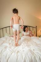 menino chinês e caucasiano de raça mista pulando na cama com sua família foto