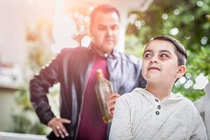 menino de raça mista com medo e machucado na frente do homem zangado segurando a garrafa de álcool foto