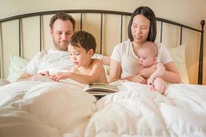 meninos chineses e caucasianos lendo um livro na cama com seus pais foto