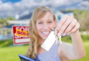 mulher segurando as chaves da casa nova com cartão em branco na frente do sinal de imóveis vendidos e casa. foto