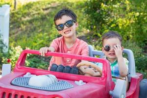 jovens irmãos chineses e caucasianos de raça mista usando óculos escuros brincando no carro de brinquedo foto