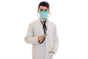 sério morena médico de uniforme com estetoscópio no pescoço e máscara posando e olhando para a câmera isolada no fundo branco foto