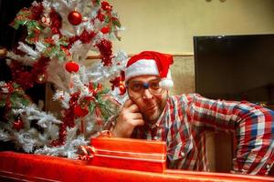 divertido nerd de óculos e chapéu de Papai Noel perto da árvore de natal foto