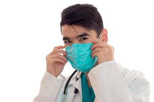 retrato do médico jovem bonito morena em uniforme branco e máscara azul com estetoscópio olhando para a câmera isolada no fundo vazio foto