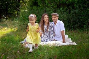 menina bonitinha de vestido amarelo esgotando seus pais. pai e mãe olhando para a filha e sorrindo no piquenique foto