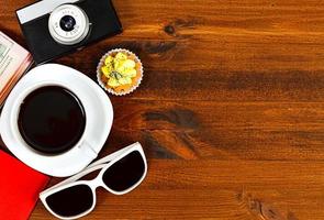 câmera fotográfica retrô, xícara de café, passaporte, óculos escuros e cupcake na mesa de madeira marrom com espaço para texto, vista superior. foto