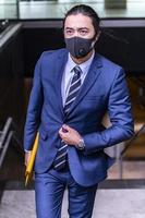 bonito empresário asiático usando máscara cobiçosa e terno caminhando ao ar livre foto
