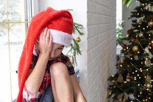 garota infeliz com um chapéu de Papai Noel senta-se no parapeito da janela de casa perto da árvore de natal. criança está chateada e chorando, esperando o natal e o ano novo. não há presentes, alegria e milagre, decepção foto