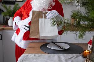 papai noel coloca na mesa da cozinha e tira sacolas de papel com presente artesanal, bolos caseiros e entrega de comida. compras, reciclagem de embalagens, feito à mão, entrega de natal e ano novo foto