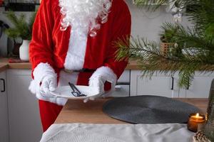 papai noel põe a mesa em casa na cozinha branca, põe prato, põe talheres garfo e faca. preparando-se para a celebração do natal, arrumando a mesa para os convidados foto