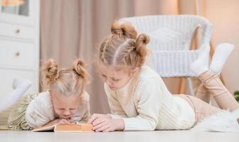 2 meninas loiras deitam no chão perto da árvore de natal e leem um livro. noites aconchegantes com um livro perto da árvore. namoradas leem um livro juntas. relacionamento caloroso foto