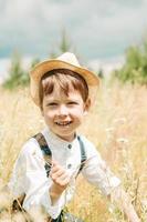 pequeno fazendeiro em um campo de verão, menino bonitinho com um chapéu de palha. menino de chapéu com uma flor fica em um campo. retrato de um menino de aldeia foto