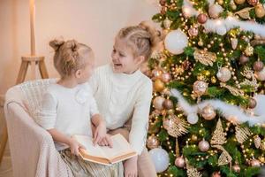 2 meninas loiras estão sentadas em uma poltrona perto da árvore de natal e lendo um livro. noites aconchegantes com um livro perto da árvore. namoradas leem um livro juntas. relacionamento caloroso foto