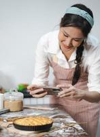 jovem usa avental tirando foto de selfie com torta caseira na cozinha. retrato de uma bela mulher asiática assando sobremesa e se divertindo tirando fotos por smartphone para redes sociais online. culinária caseira.