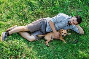 sorrindo atraente homem europeu está deitado na grama e abraçando seu cachorrinho fofo. foto