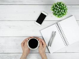 espaço de trabalho em mesa de madeira branca, espaço para cópia, caneta prateada no caderno de página em branco e tela preta do smartphome com xícara de café no conceito mínimo de duas mãos foto