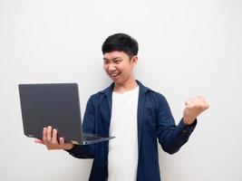 homem asiático olha para o laptop na mão e sente-se feliz, mostra o punho para cima, sorri feliz no branco isolado foto