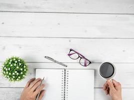 espaço de trabalho de escritório espaço de cópia de visão superior, mão com caneta prateada e xícara de café e óculos e caderno de página em branco na mesa de madeira branca foto