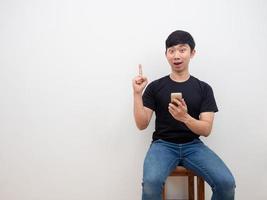 jovem asiático, segurando o smartphone na mão, tenha uma ideia, aponte o dedo para cima, sentado na cadeira fundo branco foto