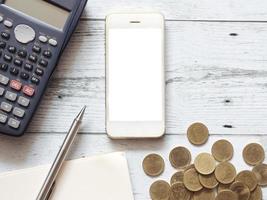 calculadora smartphone tela branca caneta prateada no caderno de página em branco e conceito de negócio de grupo de moedas de ouro, gadget de negócios para calcular na vista superior da mesa de madeira branca foto