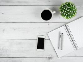 espaço de trabalho em mesa de madeira branca, espaço para cópia, caneta prateada no caderno de página em branco e tela preta do smartphome com conceito mínimo de xícara de café foto