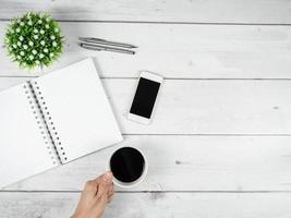 espaço de trabalho na área de trabalho de madeira branca vista de cima copie o espaço, página em branco do notebook com tela preta do smartphone e caneta prateada com mão segure a xícara de café foto